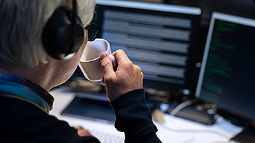 Foto på en kvinna bakifrån som sitter framför en dator och håller i en kopp kaffe.